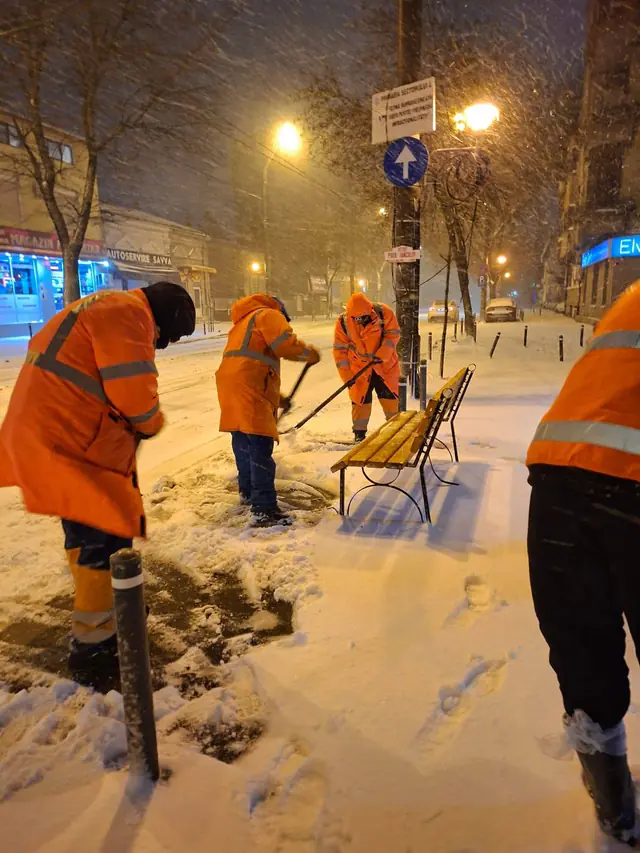 Echipe în uniforme portocalii curăță zăpada de pe un trotuar, noaptea